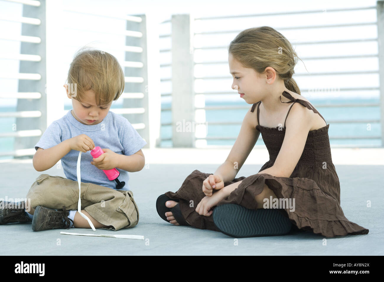 Little boy sitting on the ground inflating balloon with air pump, older sister watching Stock Photo