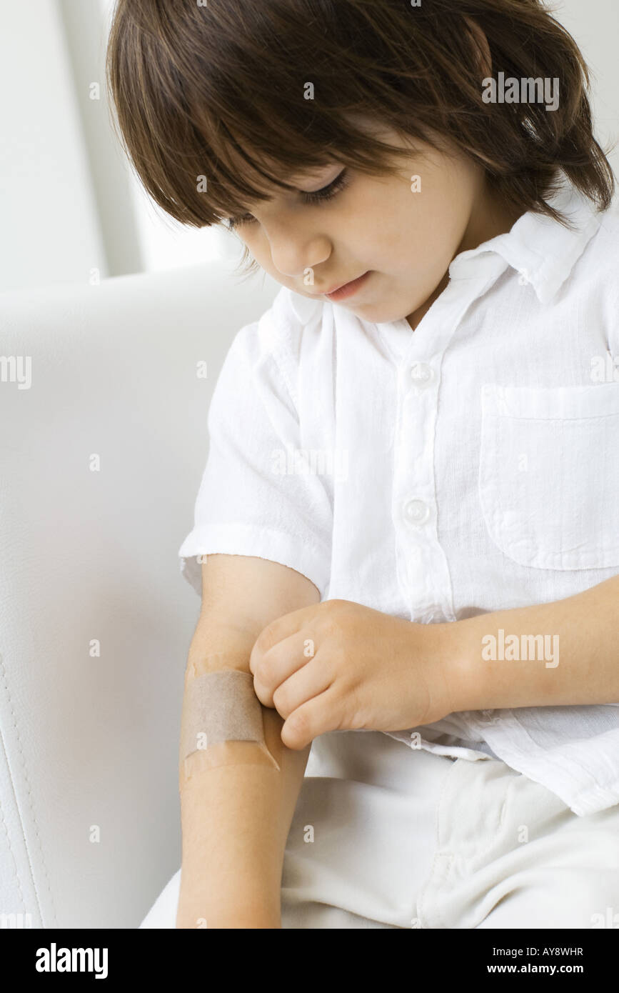 Little boy removing adhesive bandage from arm, looking down Stock Photo