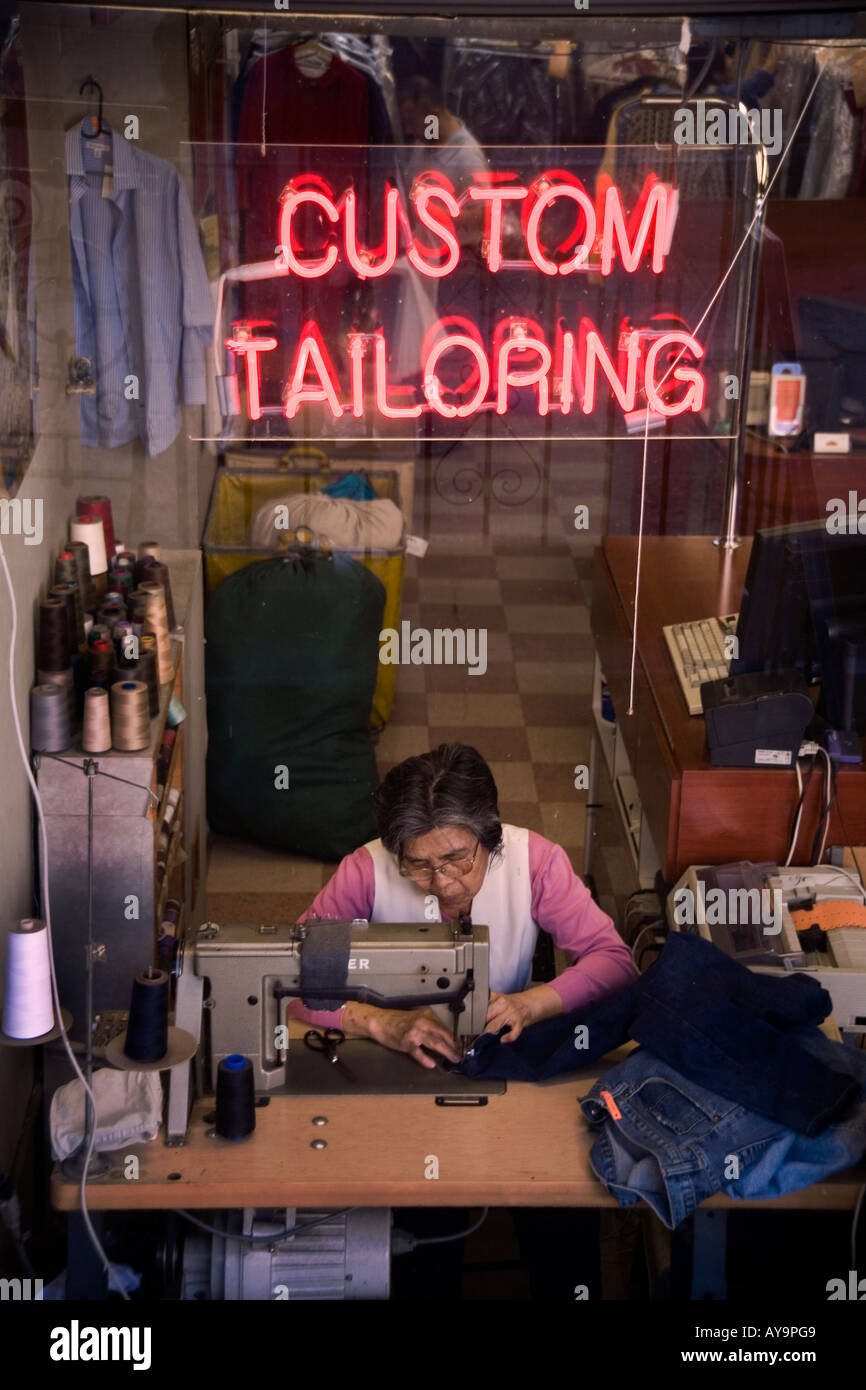 An elderly Hispanic woman works at sewing machine in the window of a custom tailor shop in New York City Stock Photo