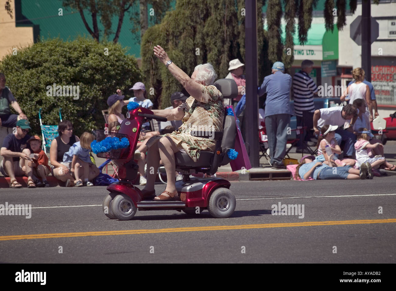 older woman riding electric wheeled chair scooter in parade waves at crowd on sidewalk Stock Photo