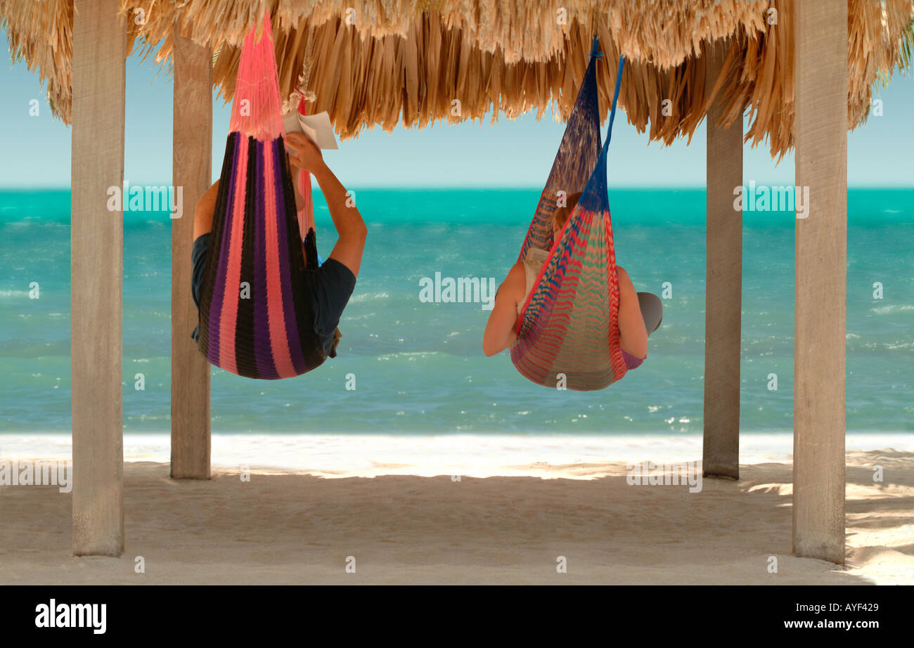 A couple relaxes in a pair of hammocks under a thatched roof on a tropical beach Stock Photo