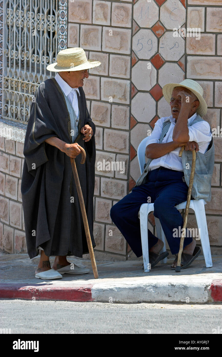 Old Berber man in traditional woollen cloak stops to talk to acquaintance at midday in shade on street Matmata Tunisia Stock Photo