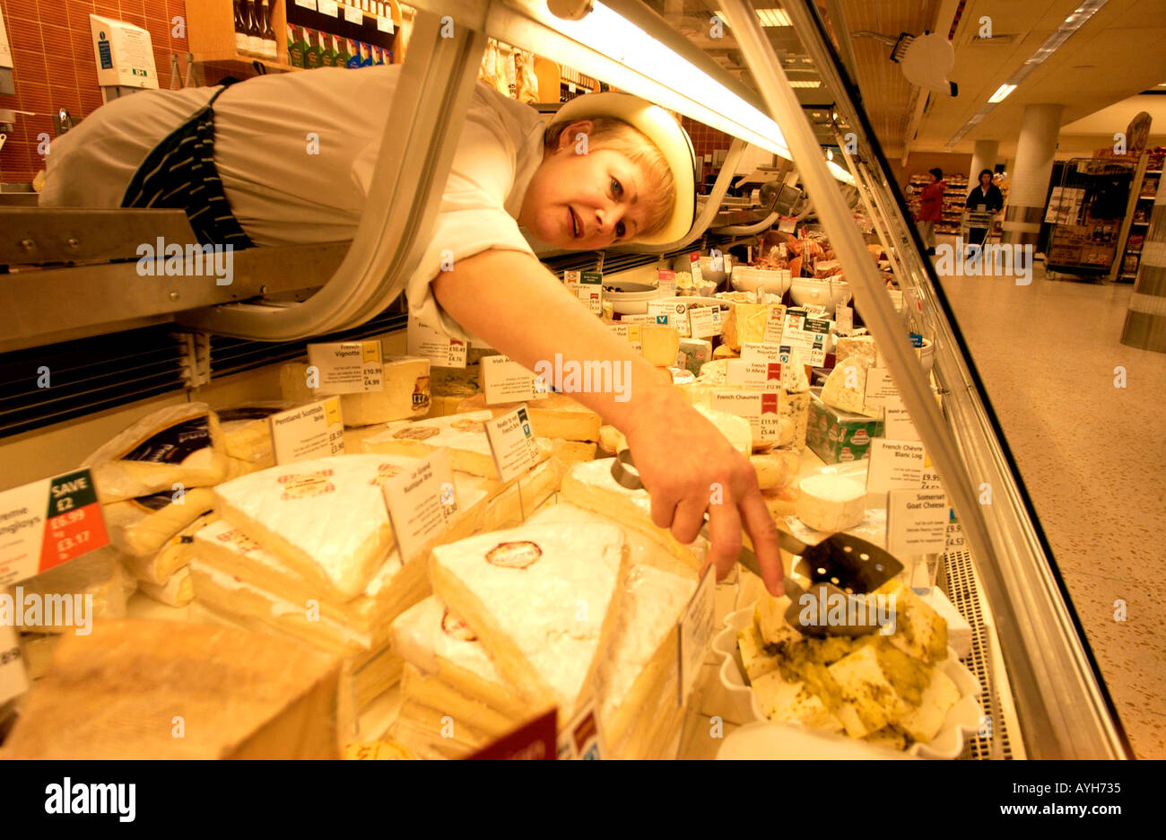 A deli assistant in uniform and hat stretches to reach cheese on her