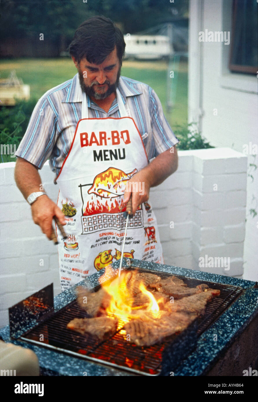 Bearded man in short sleeved shirt and amusing BBQ apron cooks steaks on flaming Bar-B-que grill in suburban garden Australia Stock Photo