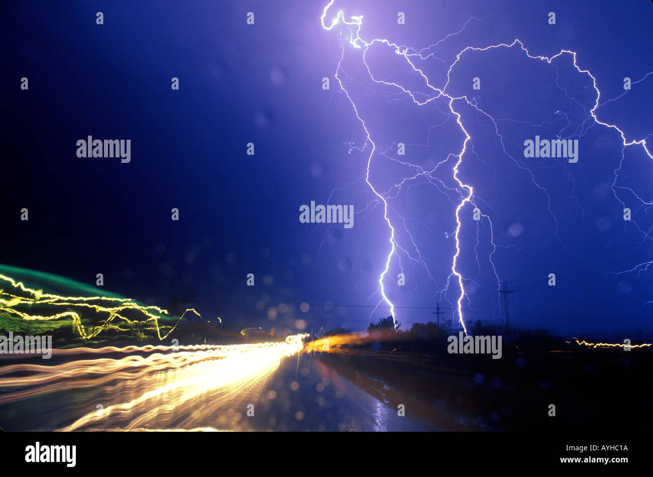 Lightning strikes near a road with car headlights blurring by. Water droplets on the windshield play in the foreground. Stock Photo