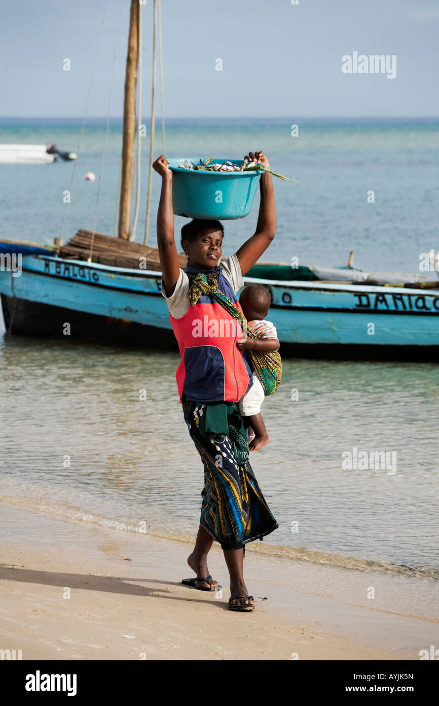 African woman with baby carrying bucket filled with crab on head Dhow sailing boat in background Benguerra Island Mozambique Stock Photo