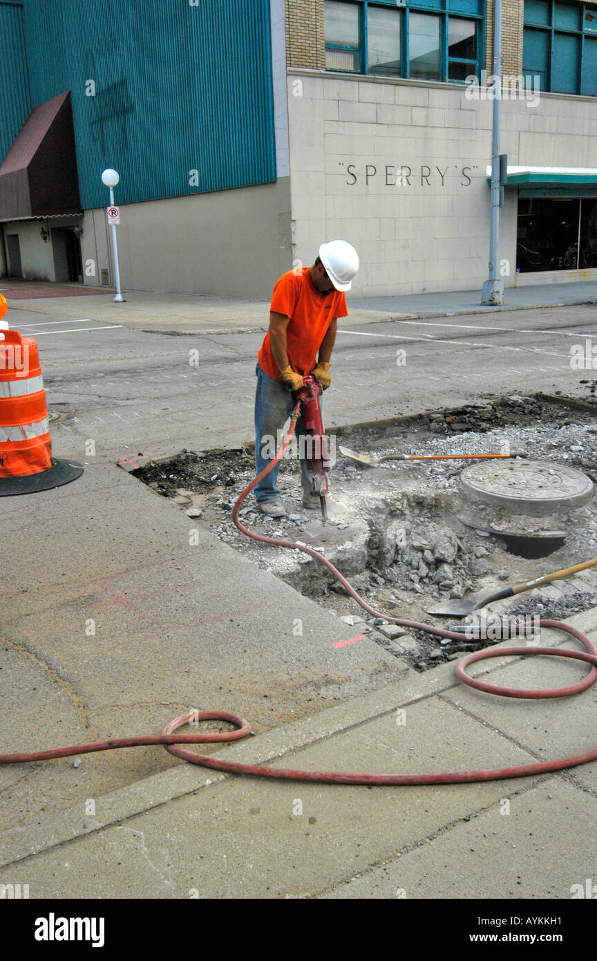 A construction worker wearing a safety helmet stands using a jackhammer to remove old cement that is under asphelt Stock Photo