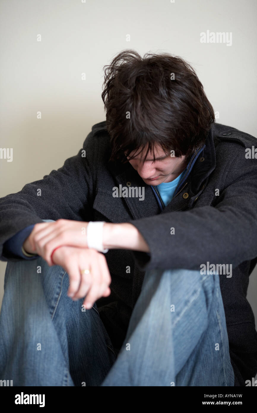 young dark haired teenage man sitting on the floor with back against the wall in the fetal position looking down Stock Photo