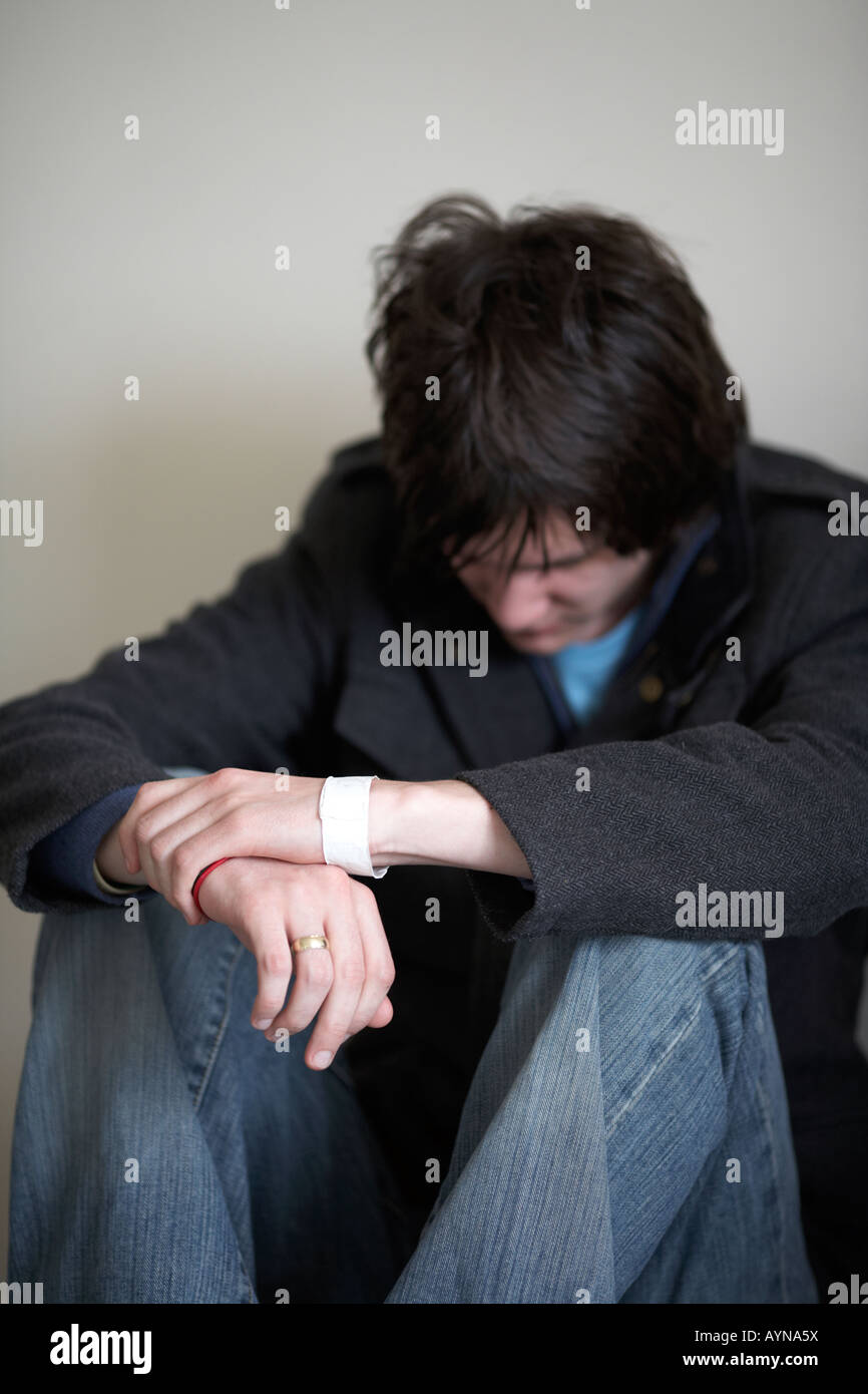 young dark haired teenage man sitting on the floor with back against the wall in the fetal position looking down Stock Photo