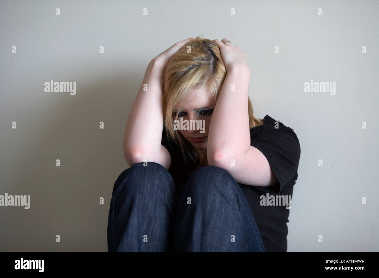 young blonde haired teenage woman sitting holding her head in her hands with her back against a wall Stock Photo