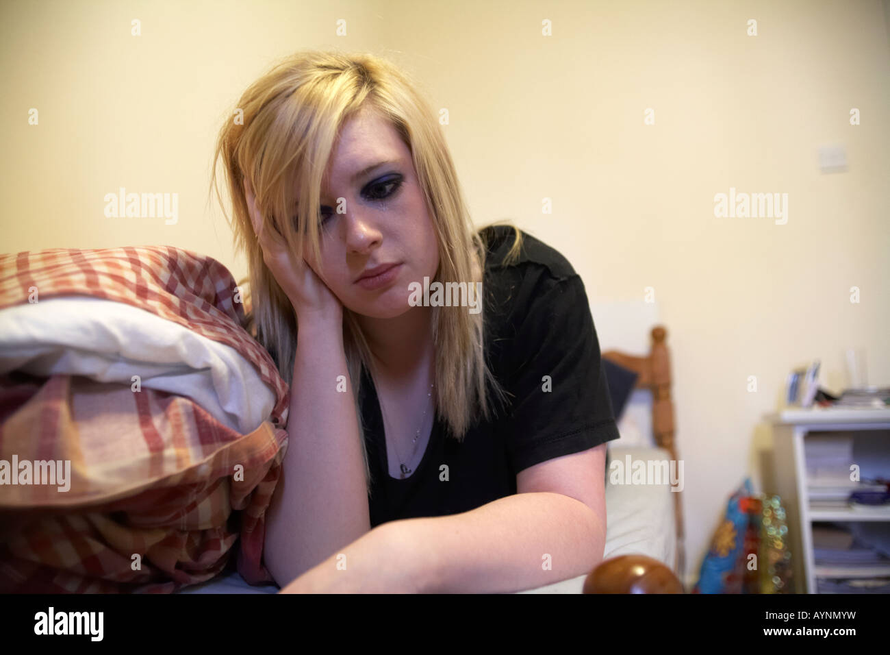 young teenage woman crying lying on bed in messy bedsit hmo bedroom holding her head in one hand Stock Photo