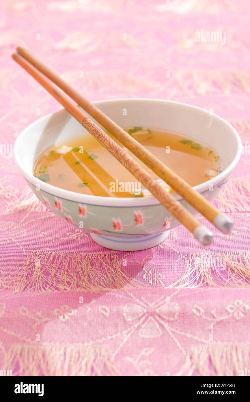 A bowl of broth and noodles sitting on a table with a pretty pink tablecloth and wooden chopsticks. Stock Photo