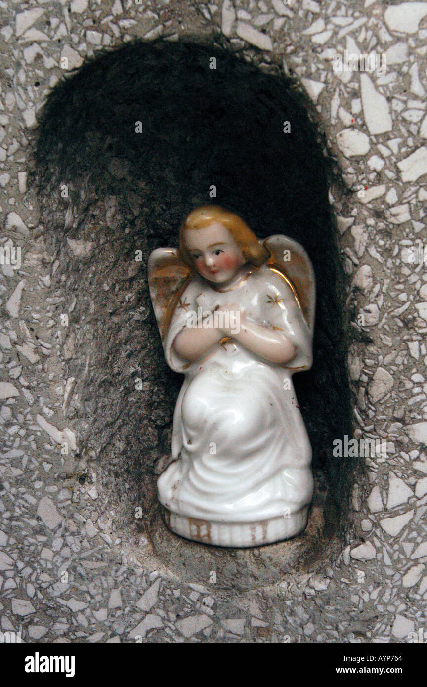 Small statue of angel in a dark niche of a textured stone wall. Porcelain figurine of a praying angel with gold-edged wings and blonde hair. Stock Photo