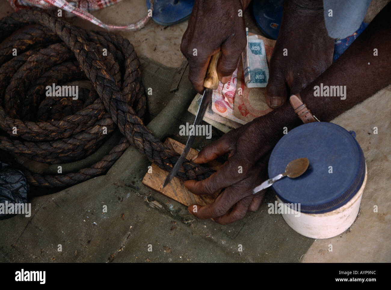 piece of tobacco NIGERIA West Africa Benue Vendor cutting piece of tobacco from plaited coil  at stall in market with paper money on ground Stock Photo