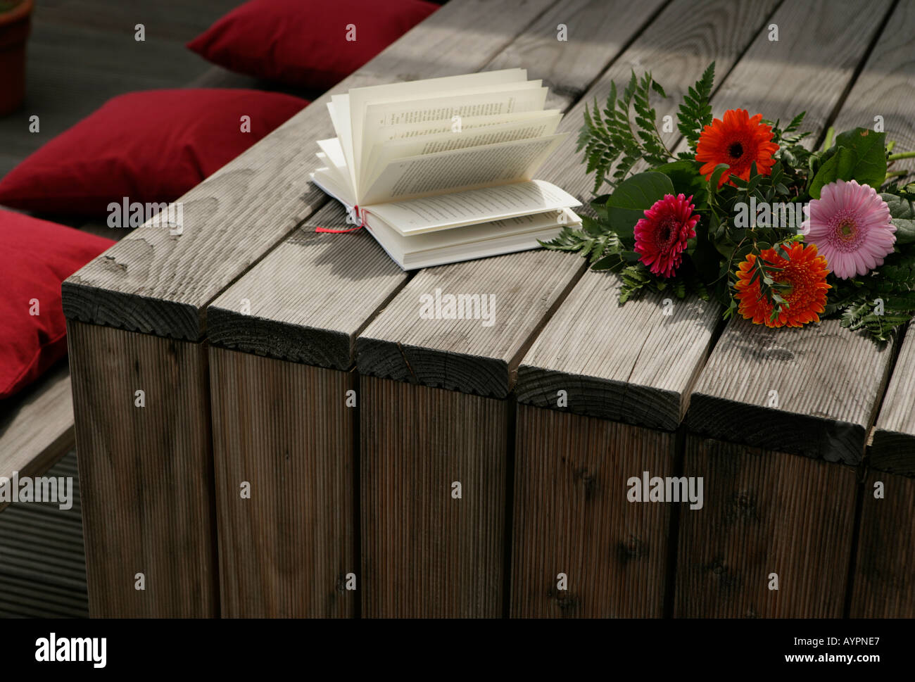 Bunch of flowers and a novel seen on a wooden table with some red cushions beside it Stock Photo