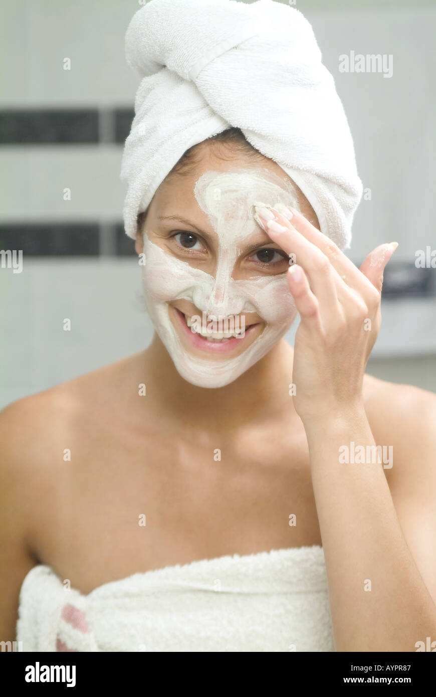 A young woman applies cream on her face as she smiles at the camera Stock Photo