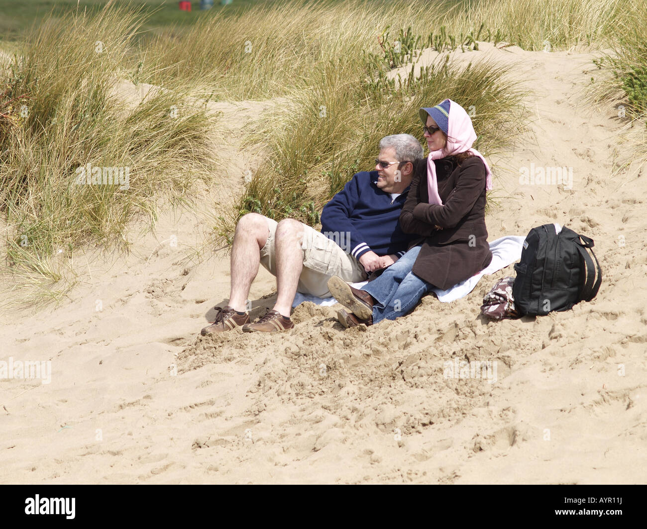Older couple sitting on sand dunes at Summerleaze Beach, Cornwall, on a windy spring day. Stock Photo