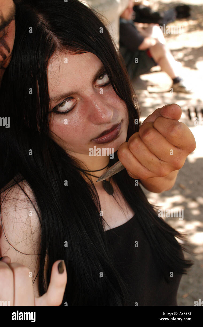 Portrait of a Satanist woman with long black hair and white contact lenses, featuring dark makeup and a knife held near her face. Stock Photo