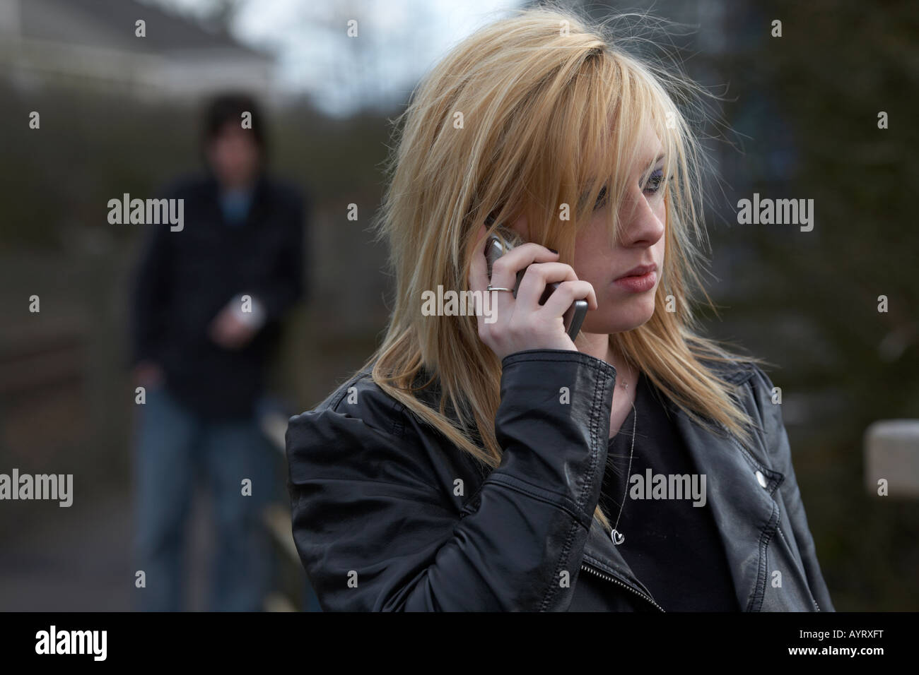 blonde haired teenage woman on mobile phone making a call standing with man standing watching close behind woman being stalked social distancing Stock Photo