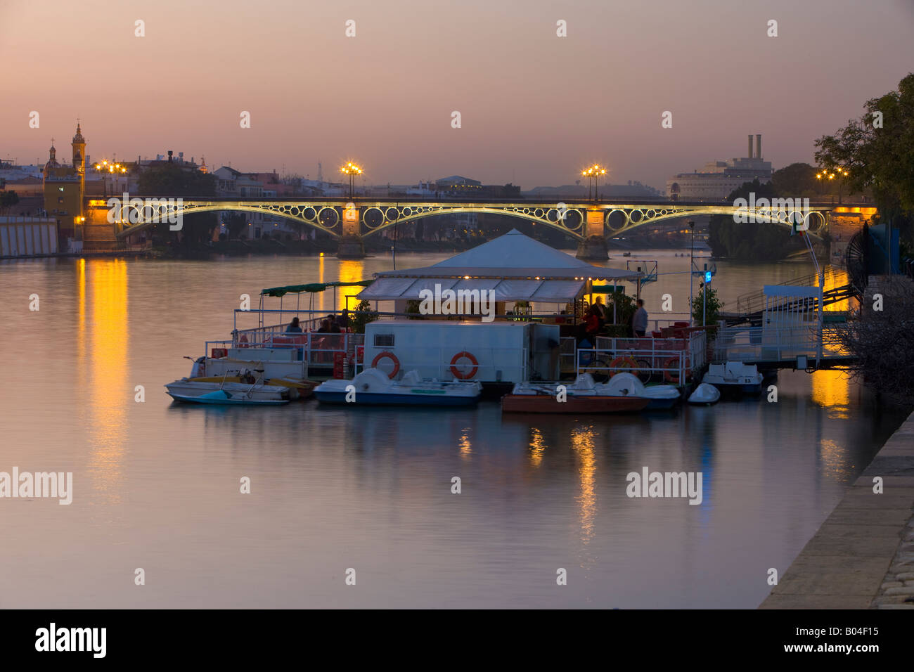 Puente de Isabel II across Rio Guadalquivir (River) to Triana district with boat dock and restaurant in the foreground at dusk Stock Photo