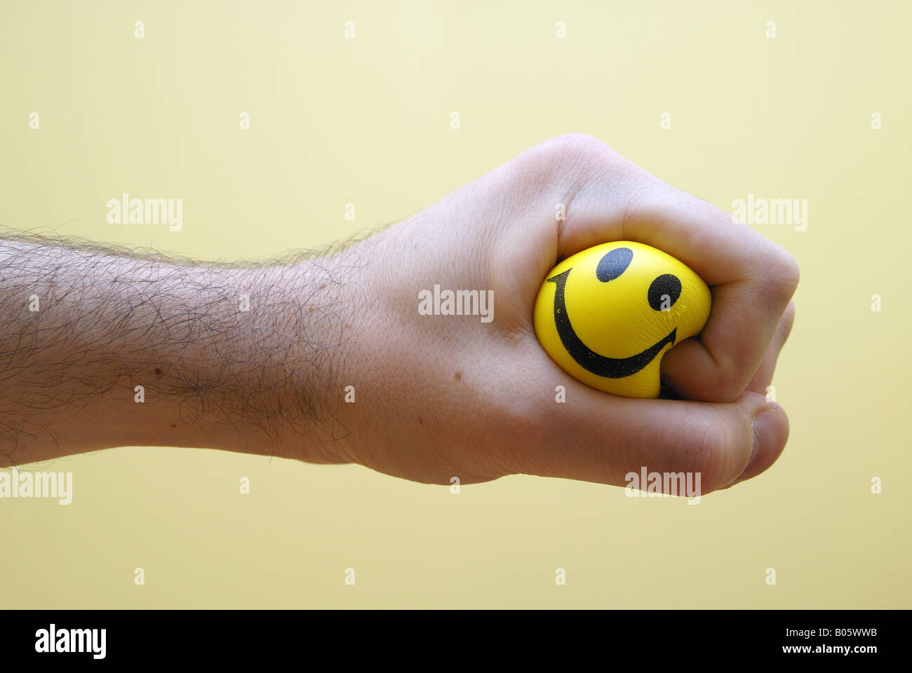 A hand clenches a rubber stress ball.  A happy face printed on the ball can be seen between the fingers of the hand. Stock Photo