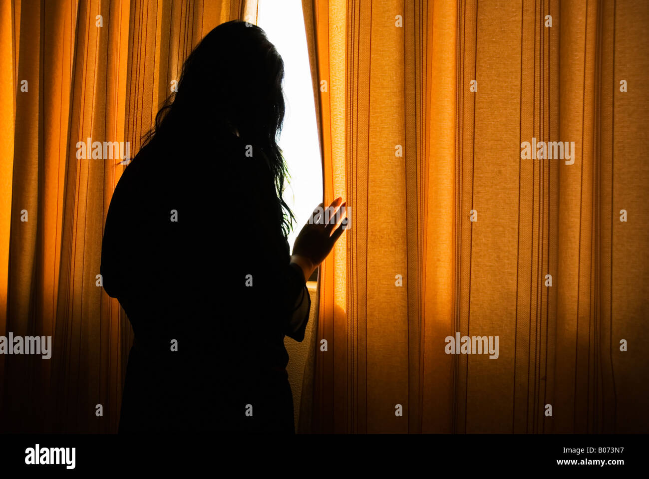 Mystery woman is looking through a gap in the curtains.She may be suffering from an illness such as depresion or agoraphobia. Stock Photo