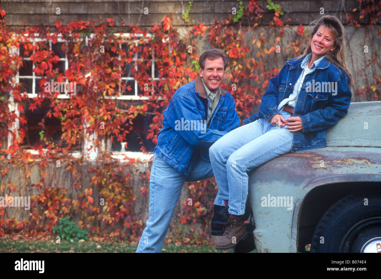 A smiling man and woman their 30s or early 40s wearing blue denim attire enjoys an old grey pickup truck in autumn weather. Stock Photo