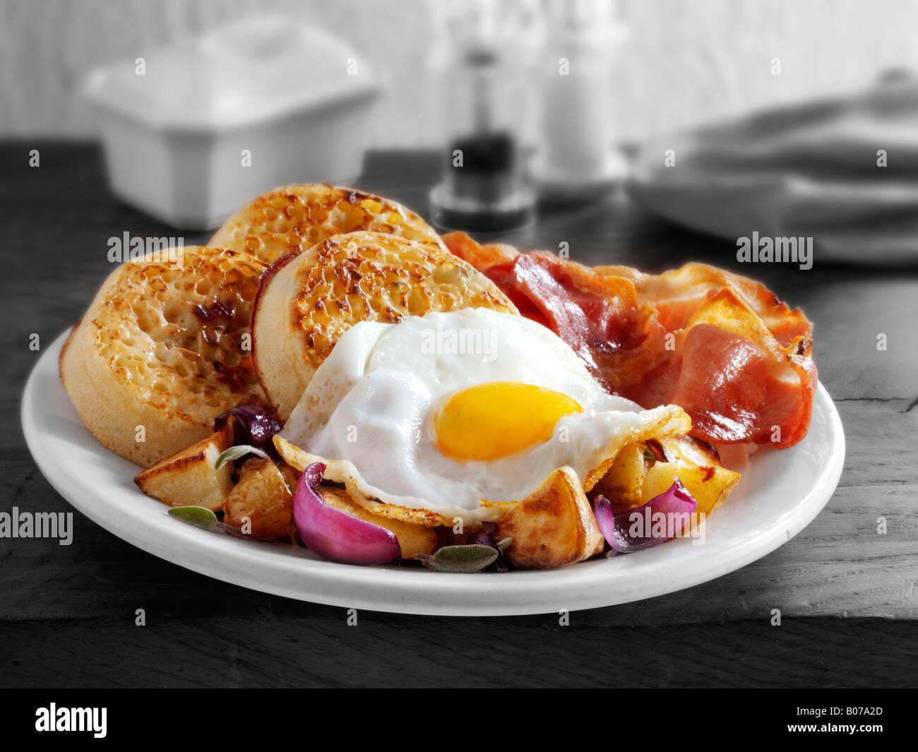Full English Breakfast with crumpets, served on a white plate in a table setting - Fried egg, bacon, sautéed potatoes and crumpets Stock Photo