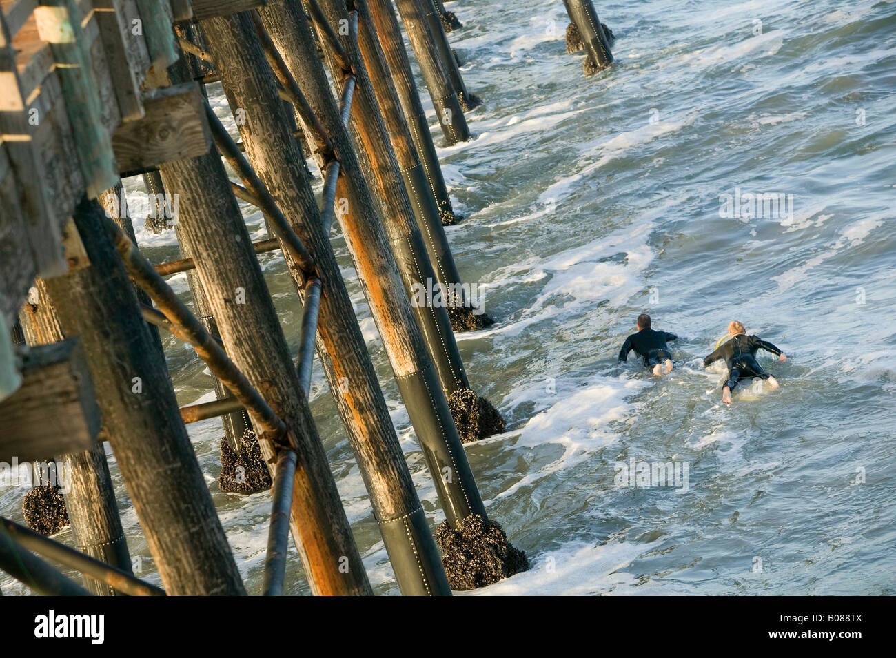 Two surfers paddle their way past a pier for a morning of surfing. Stock Photo