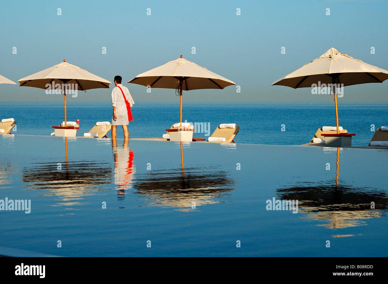 Tourist walking between deck chairs and shade umbrellas reflected in the Chedi Hotel swimming pool with a view over the sea, Mu Stock Photo