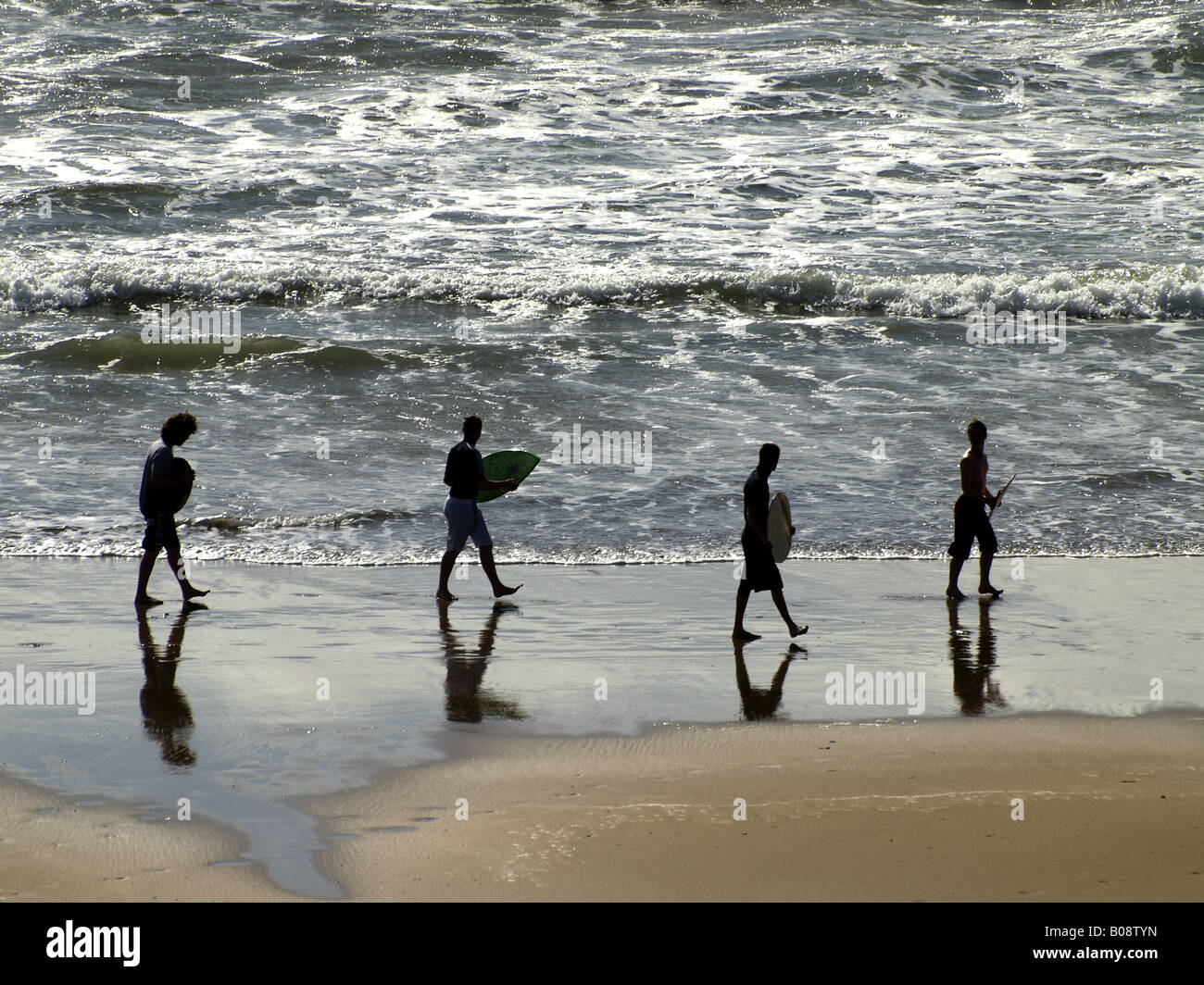 Silhouette of four young men with skimboards walking along the waters edge at sunset. Stock Photo