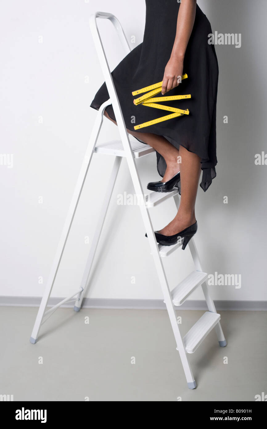 Dark-skinned woman wearing a black dress and high heels standing on a ladder, holding a yardstick Stock Photo