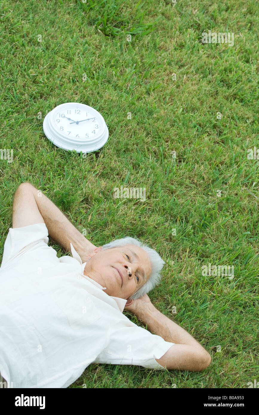 Senior man lying on grass next to clock, hands behind head, high angle