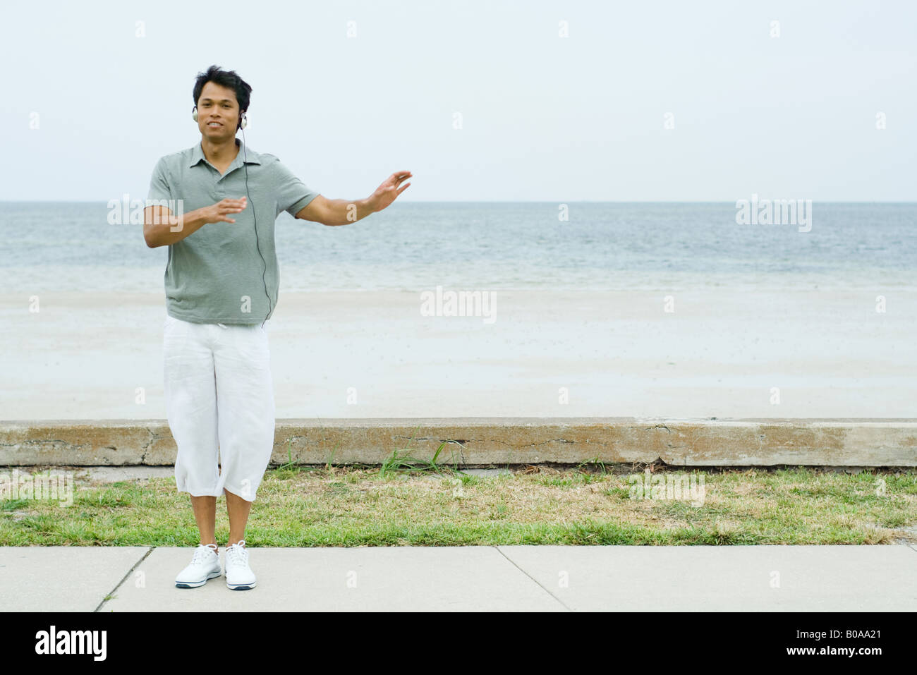 Man standing at the beach with arms out, listening to headphones, smiling at camera Stock Photo