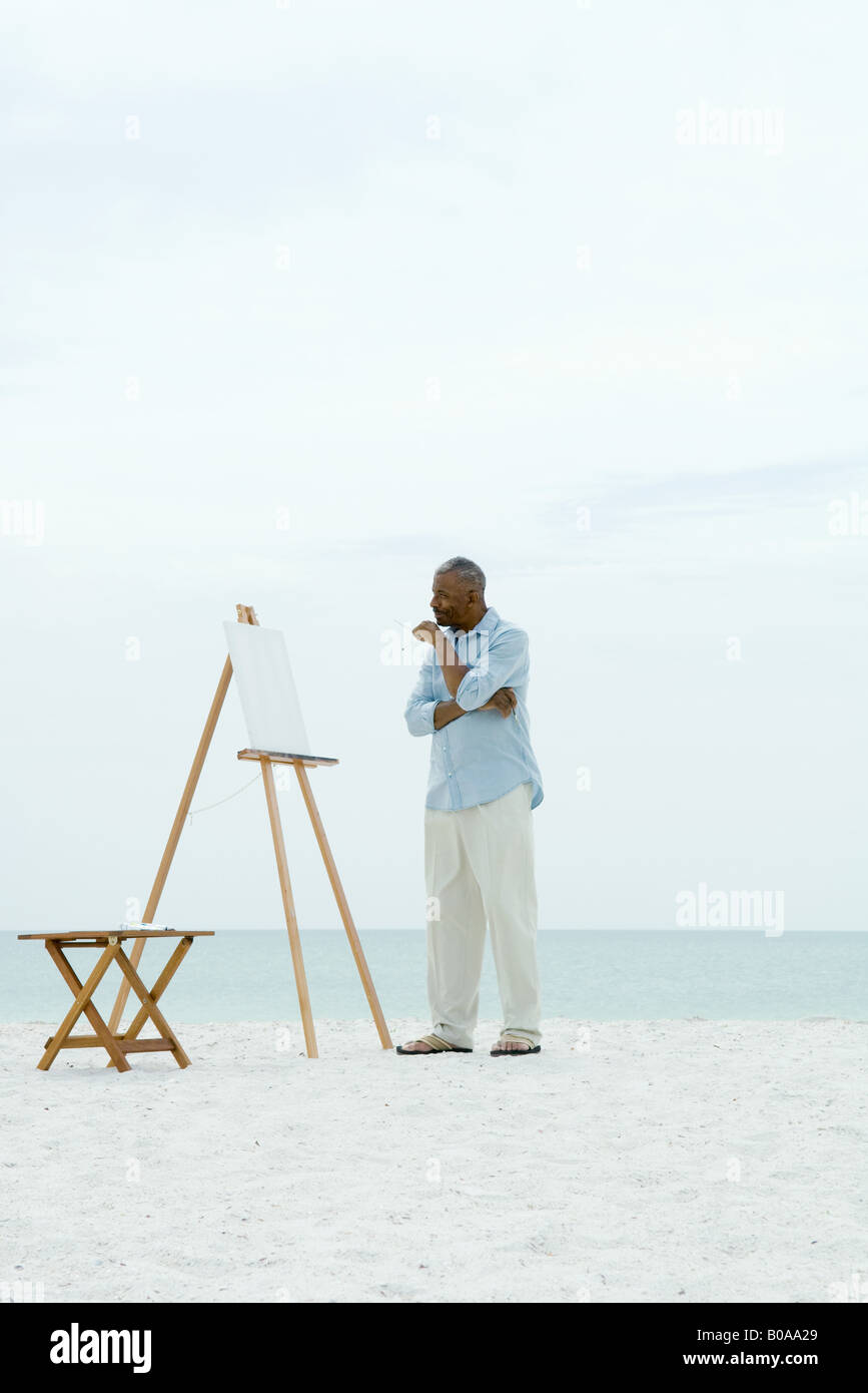 Senior man standing in front of blank canvas at the beach holding