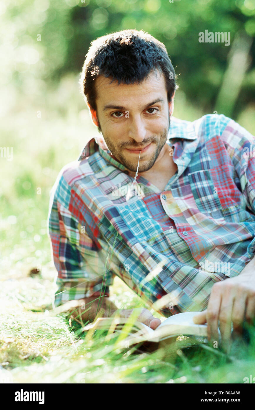 Man reading book outdoors, smiling at camera, blade of grass in mouth Stock Photo