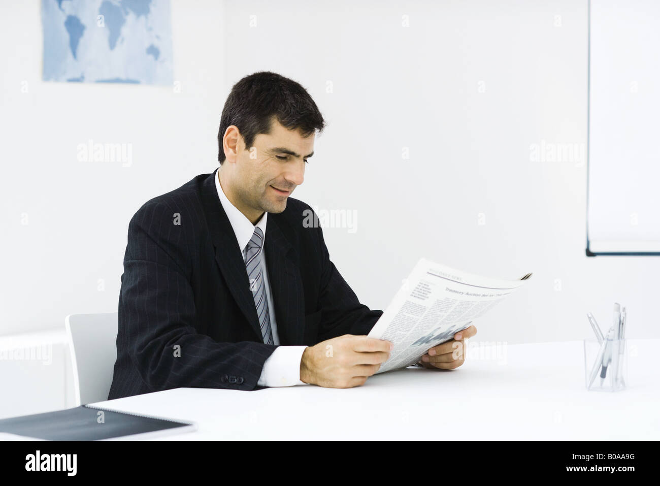 Businessman sitting at desk, reading newspaper, smiling Stock Photo