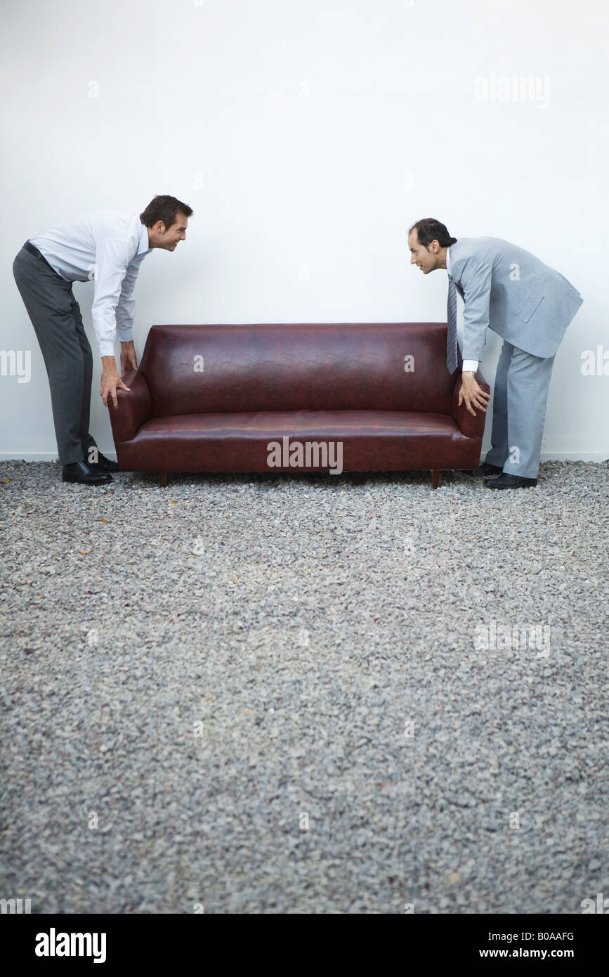 Two businessmen standing picking up sofa, looking at each other, side