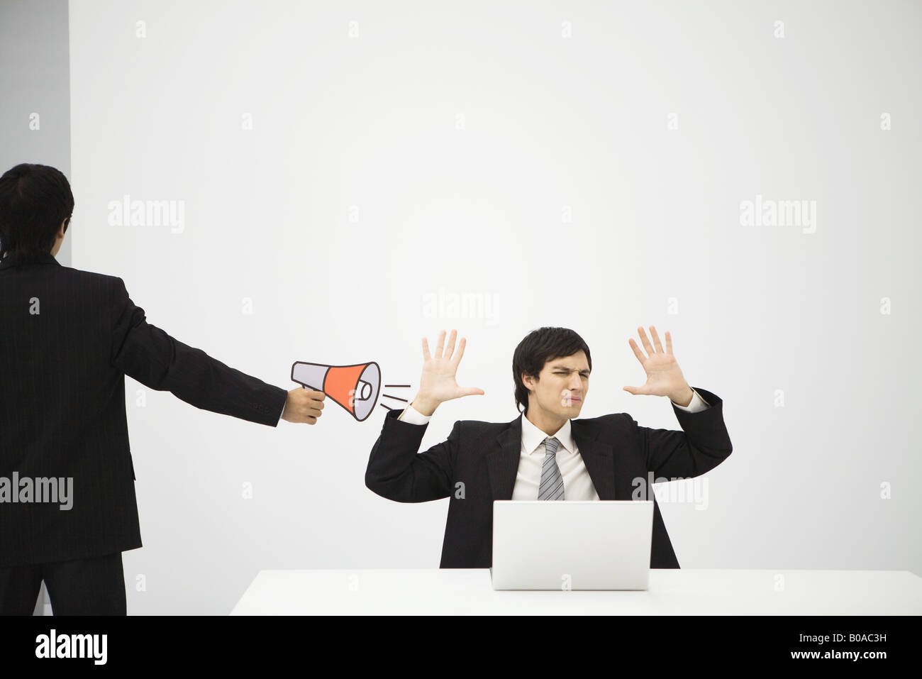Young professional sitting in front of laptop computer with hands raised, colleague aiming megaphone at him Stock Photo