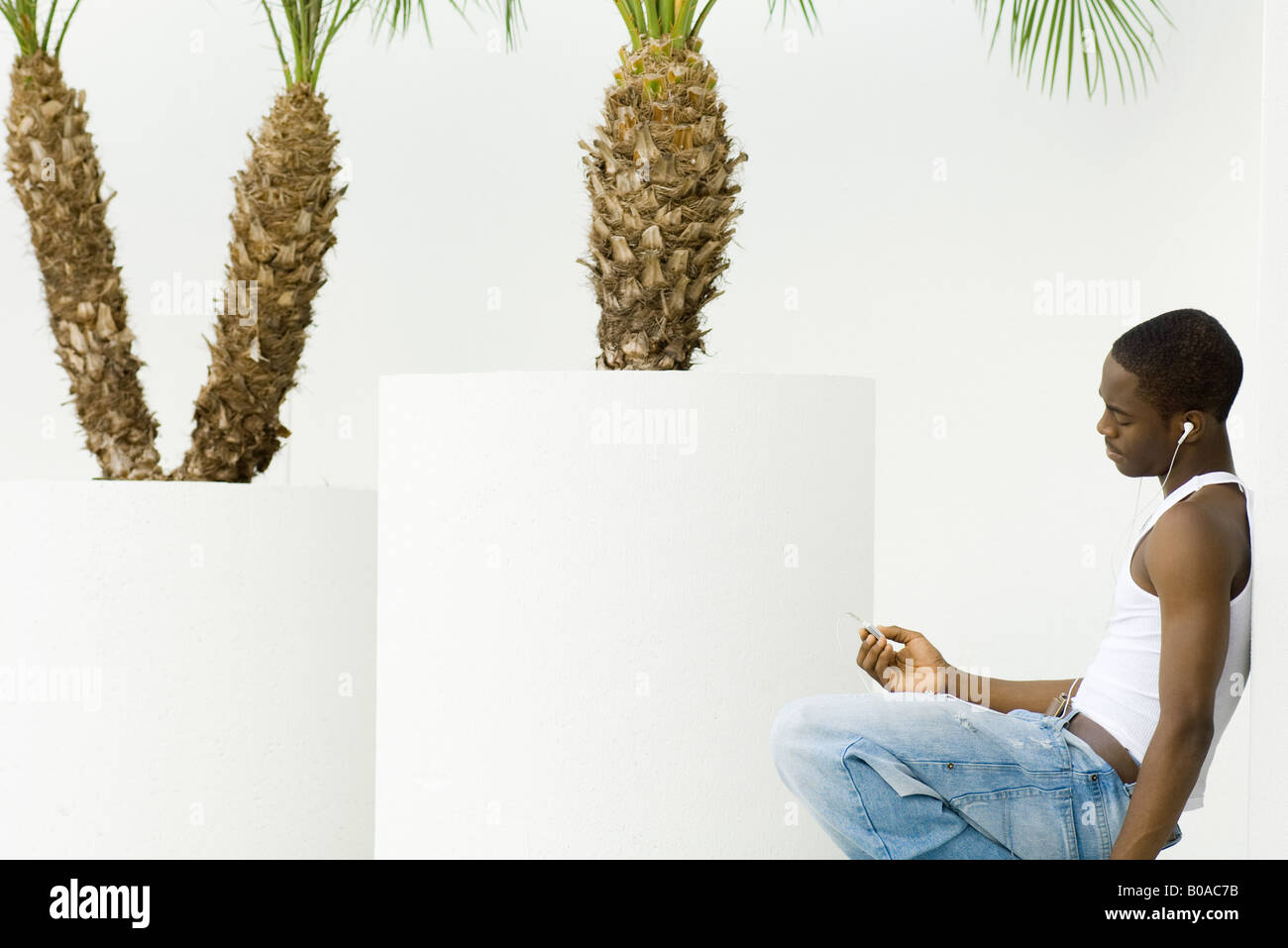 Teen boy listening to MP3 player, crouching, leaning against wall, potted palm trees in background Stock Photo