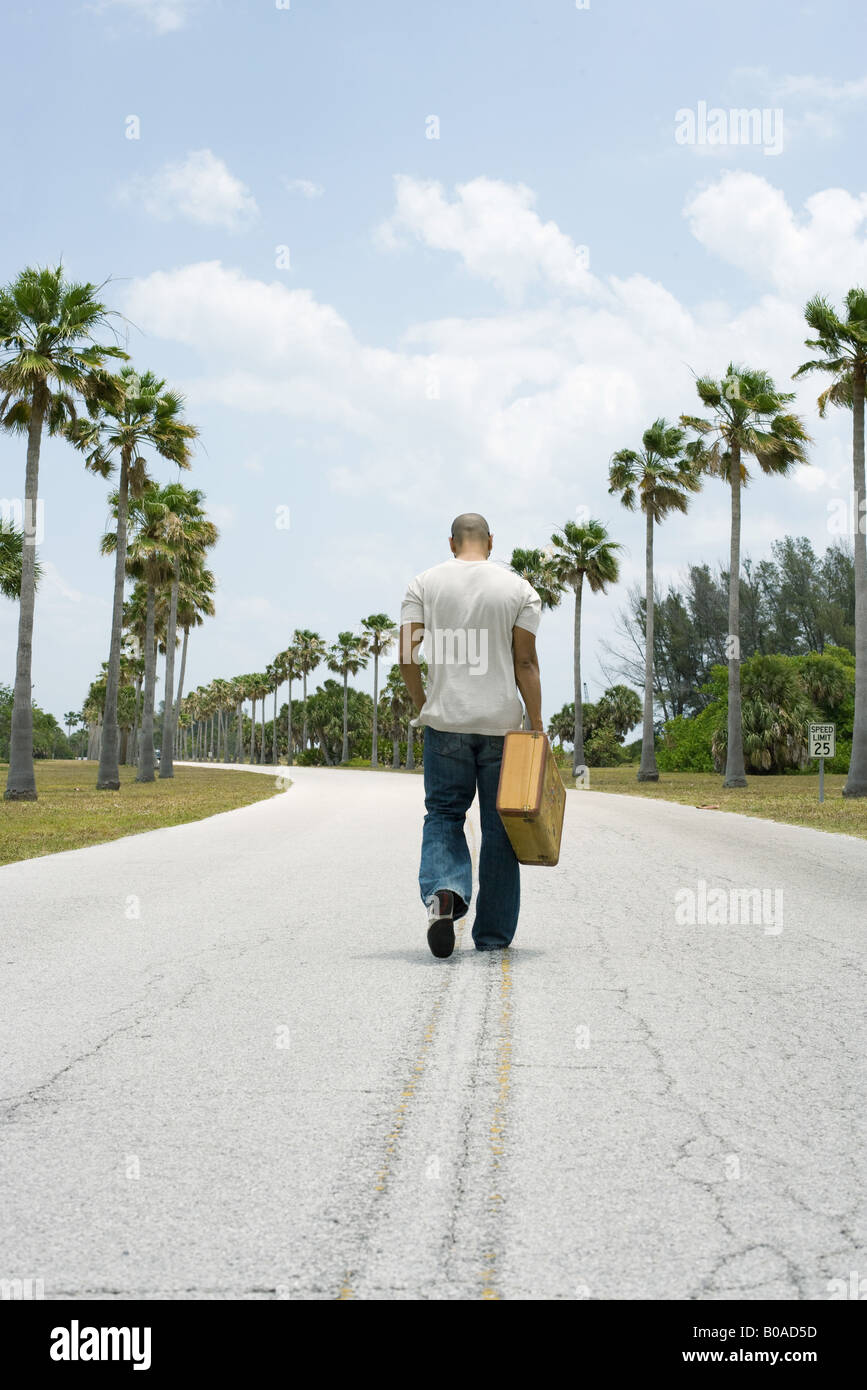 Man walking in center of road, carrying suitcase, rear view Stock Photo