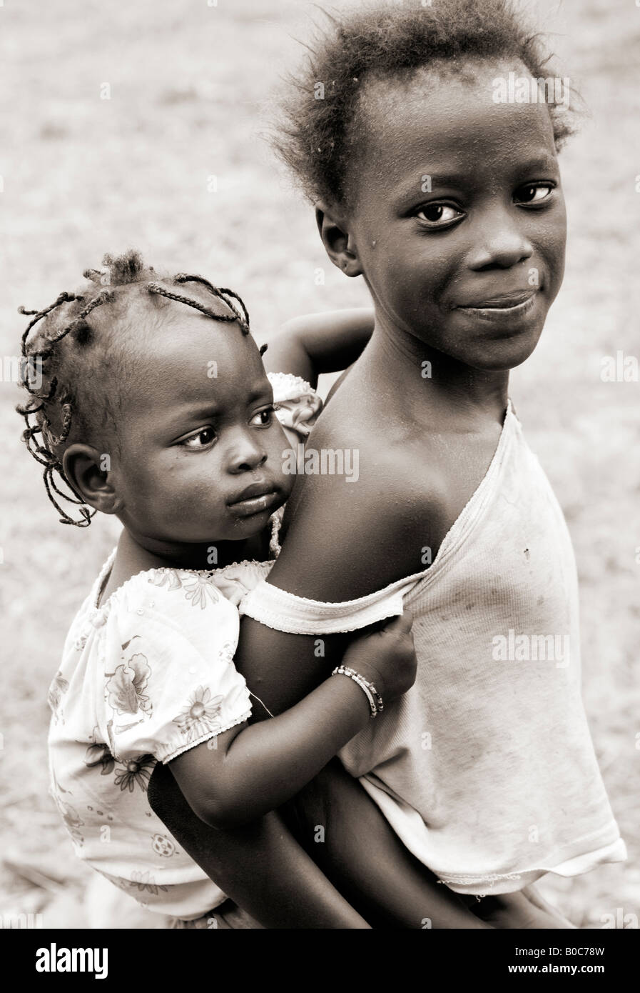 Black and white, monochrome, sepia toned photograph of a young Gambian African girl carrying her baby sister piggy-back style. Stock Photo