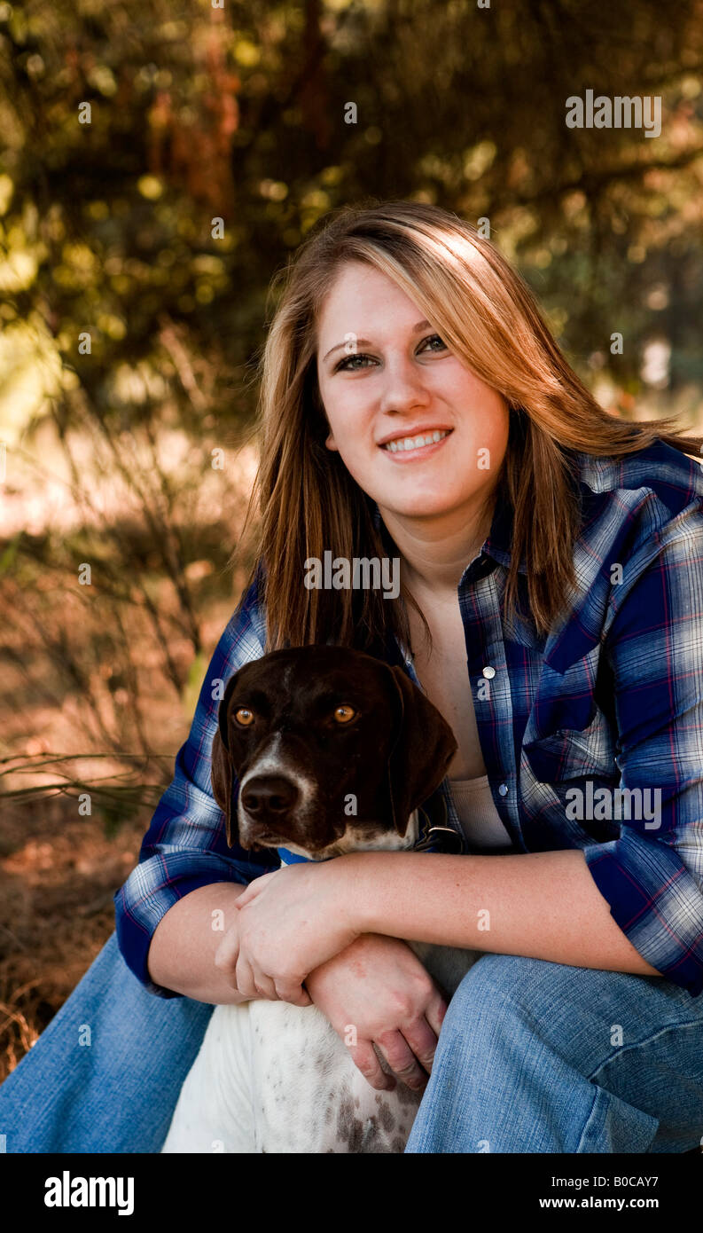 Image of a blonde teenaged girl dressed in a blue white flannel shirt and jeans sitting with her arms around a hunting bird dog Stock Photo