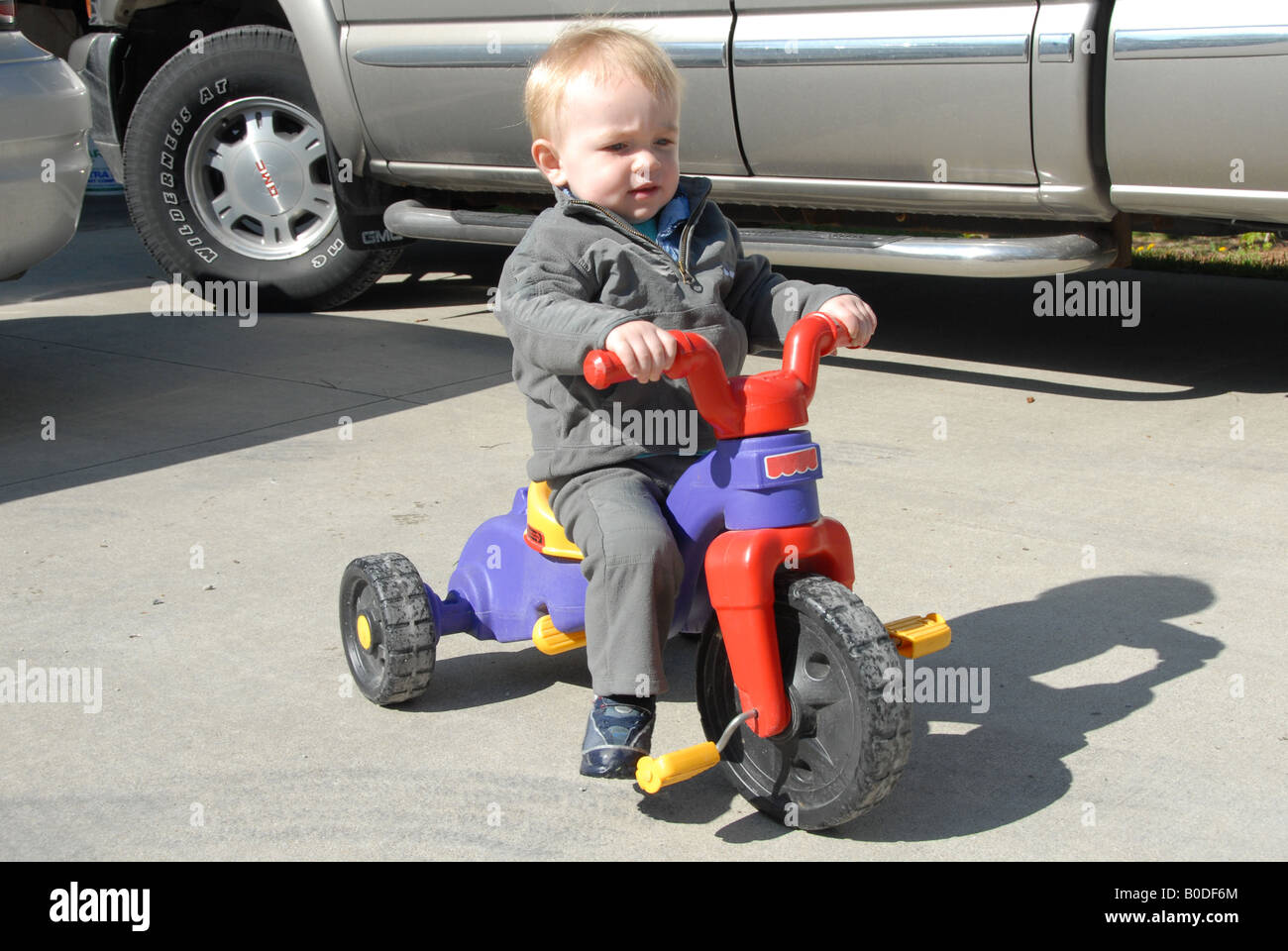A toddler boy rides a tricycle outside on the pavement on a sunny day. Stock Photo
