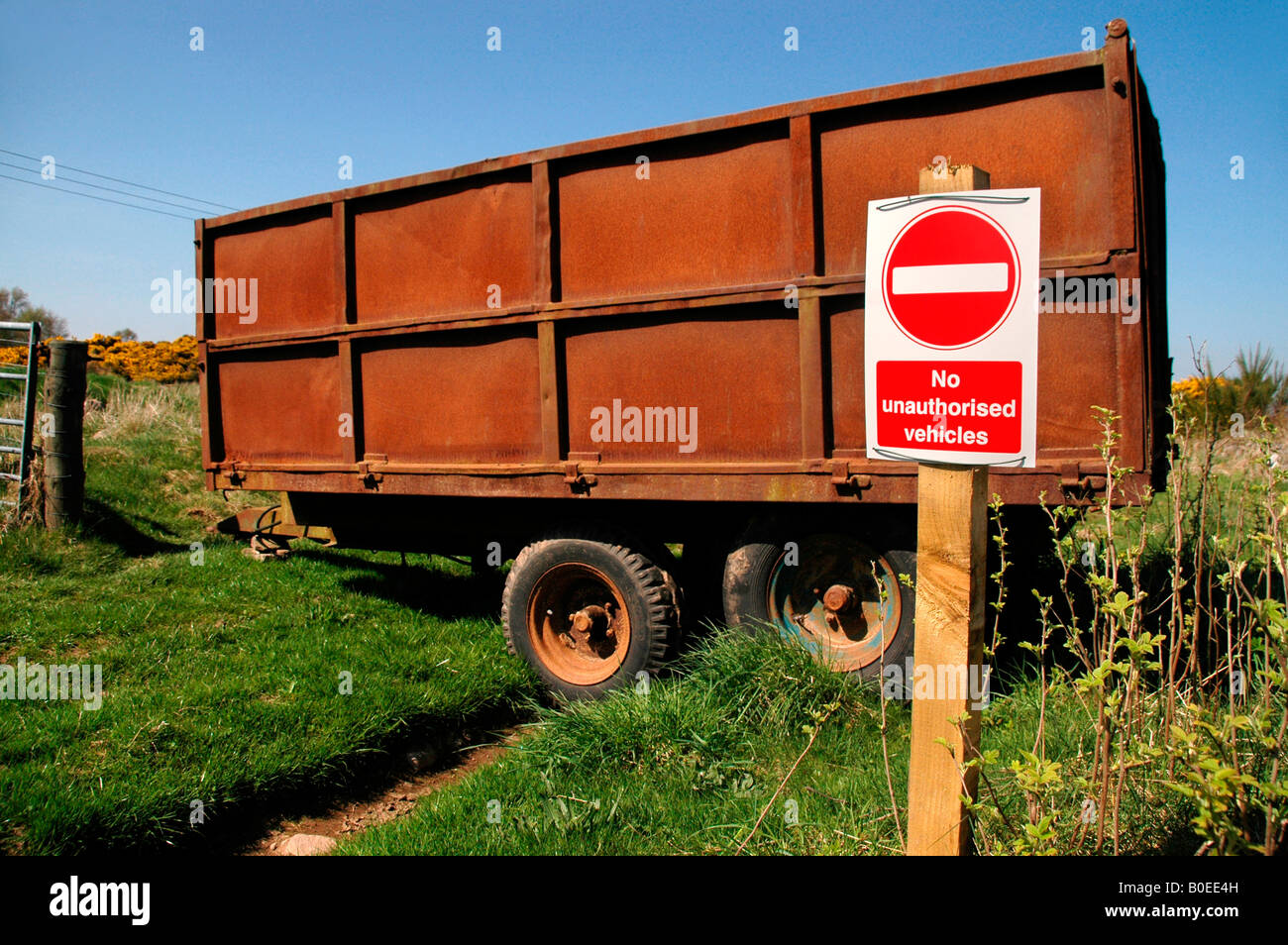 A 'no entry sign' with the caption - 'No unauthorized vehicles', with a rusty farm trailer parked across the track. Stock Photo