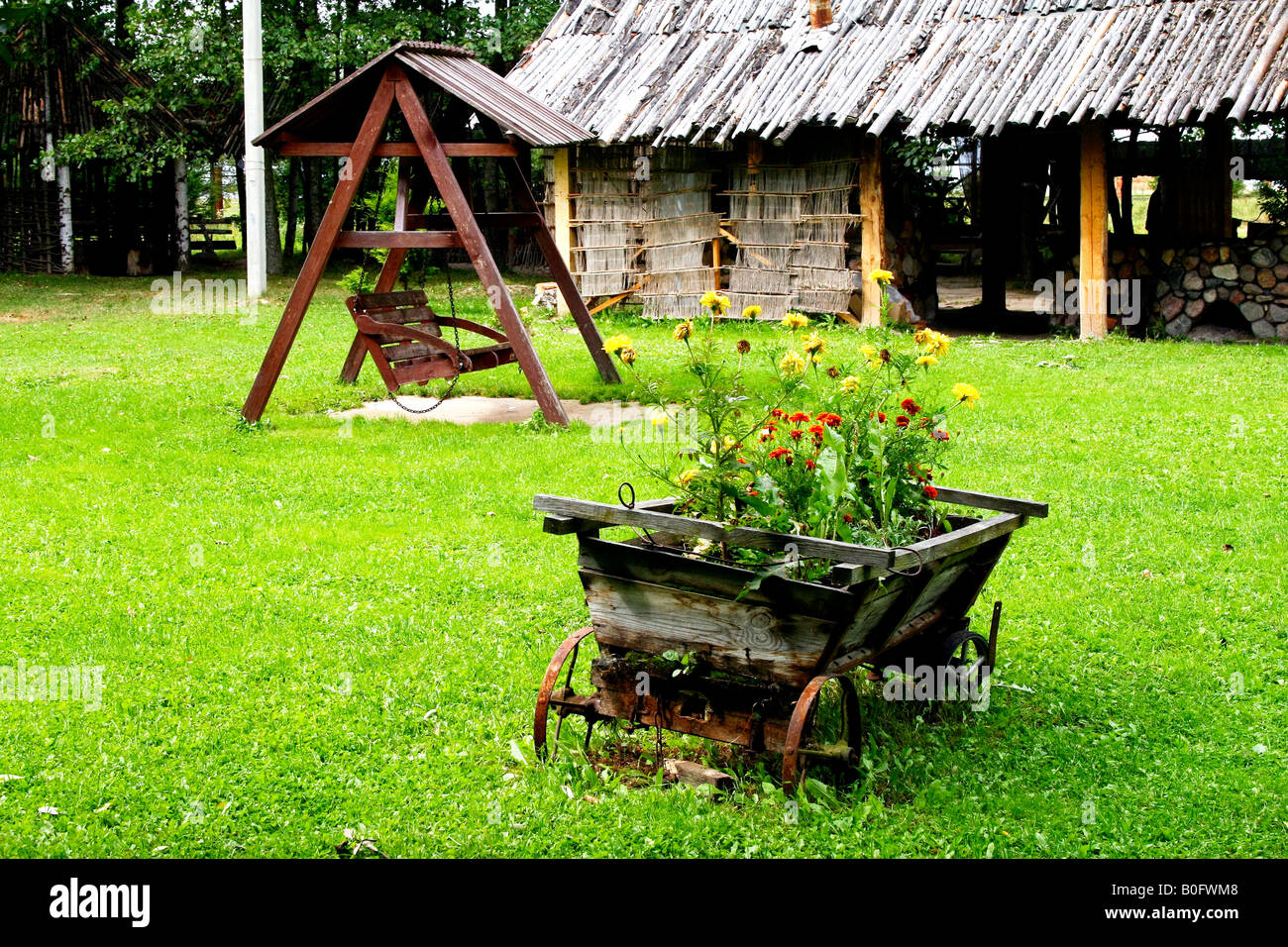 Flower Carriage (cart), swing and a wooden hut at grassy meadow. Dudutki Village, Belarus, September 2007 Stock Photo