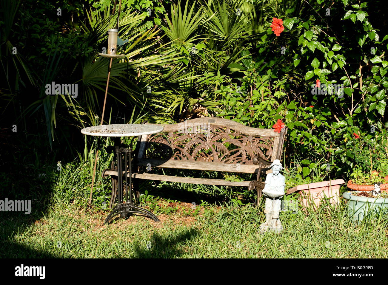 Bench with bird feeder over a table with red hibiscus flowers with a small statue with a creeping shadow in Florida Stock Photo