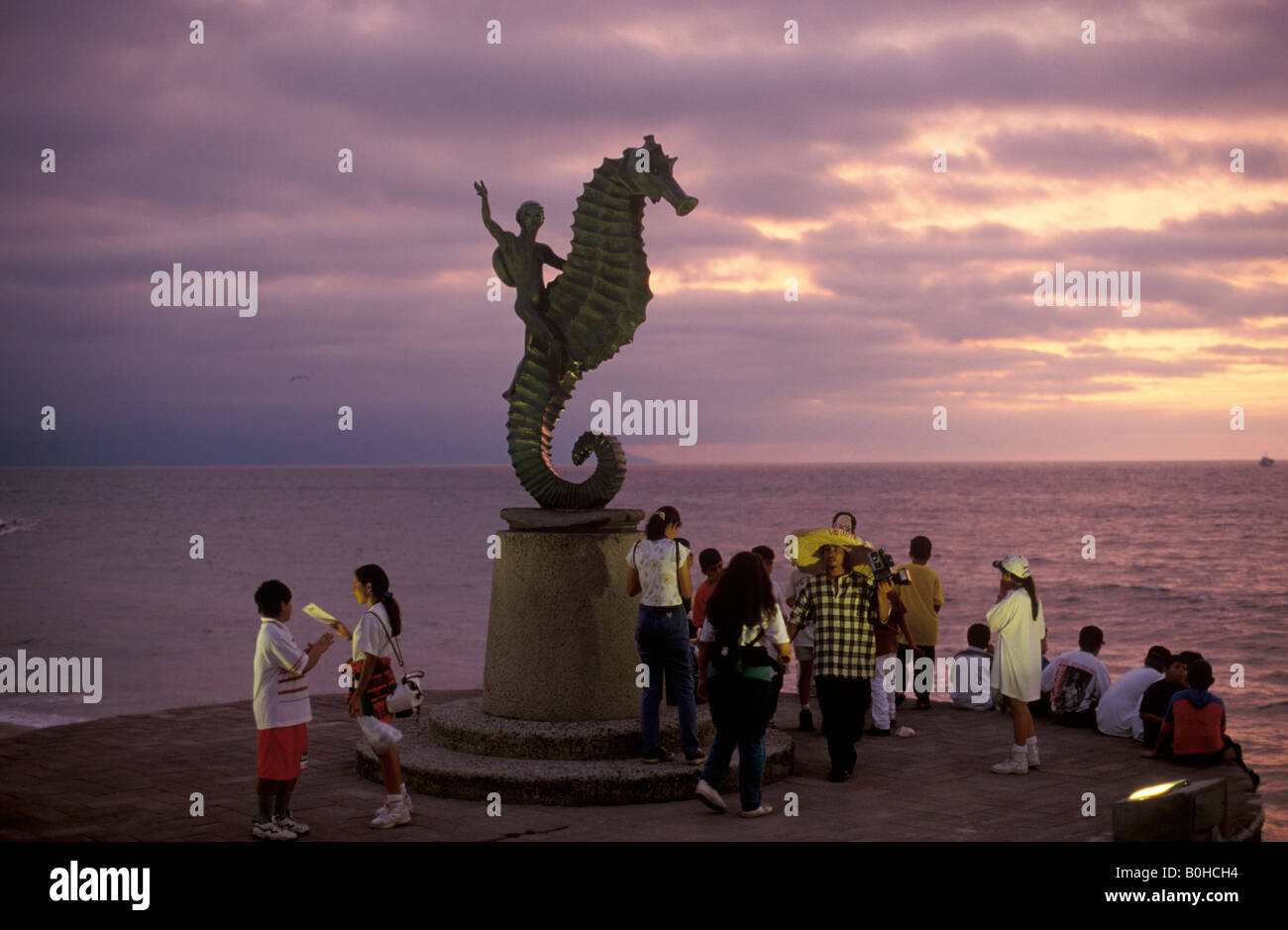 Tourists enjoying the sunset view beneath a statue of a person riding a seahorse on the beach promenade in Puerto Vallarta, Jal Stock Photo