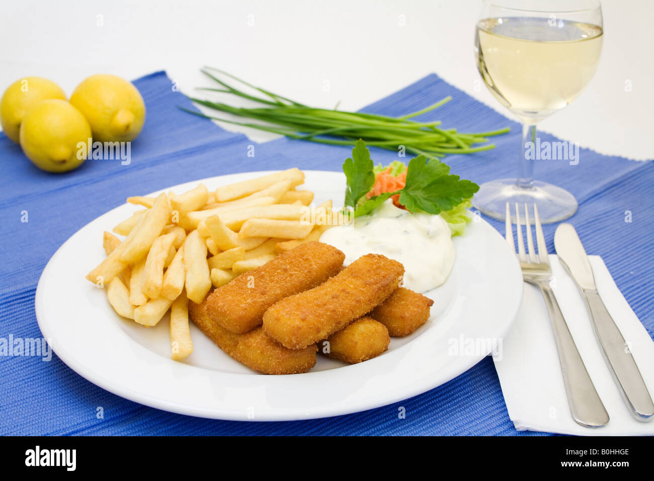 Fish sticks, tartar sauce and french fries served with a glass of white wine, lemons and chives Stock Photo