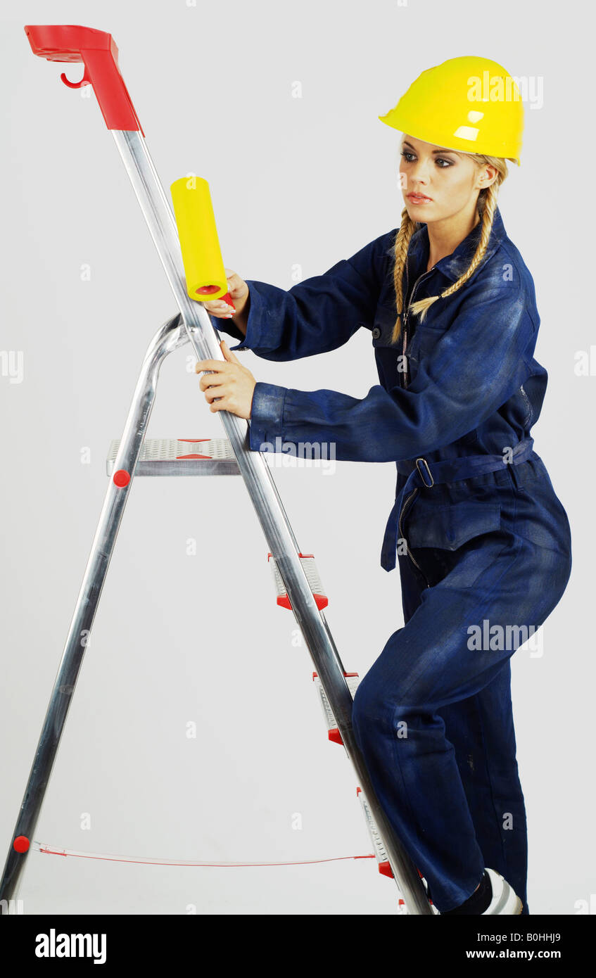 Young woman, construction worker wearing yellow hardhat on a ladder with a paint roller Stock Photo